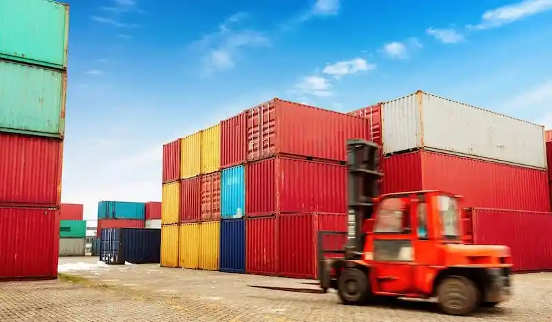 Red forklift moving quickly past large stacks of multi-colored shipping containers under a bright sky.
