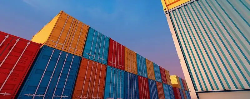 Low-angle view of colorful stacked shipping containers (red, yellow, blue) under a sunset sky, symbolizing vast inventory.