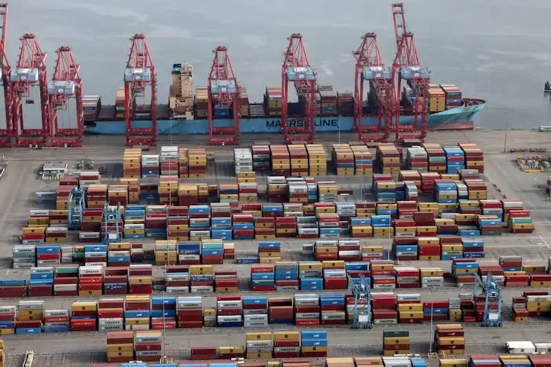 Aerial view of a vast container port, loaded cargo ship, and many red gantry cranes, symbolizing global trade volume.