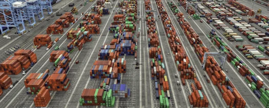 High-angle aerial photo of a vast, organized intermodal container terminal, showing hundreds of stacked red, blue, and green shipping containers arranged in long rows.