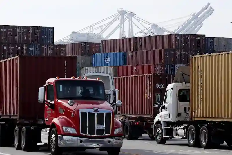 Red and white semi-trucks hauling containers, driving past stacked containers with enormous gantry cranes in the background.