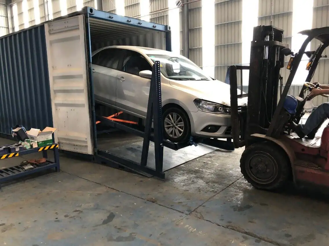 Forklift assisting in loading a silver sedan onto a specialized steel ramp system inside an open shipping container.