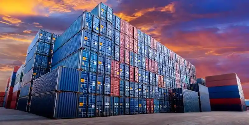Dramatic sunset photo of towering stacks of blue and red shipping containers, symbolizing the massive weight in logistics.