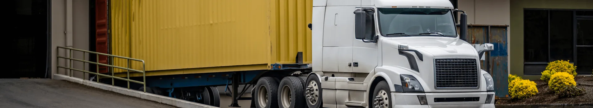 A clean, white semi-truck backing a bright yellow shipping container up a ramp toward a loading dock at a commercial or industrial facility.