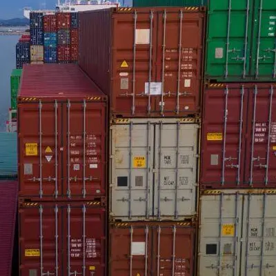 Aerial side view of containers stacked high on a cargo ship, passing a large port terminal, symbolizing transport cost.