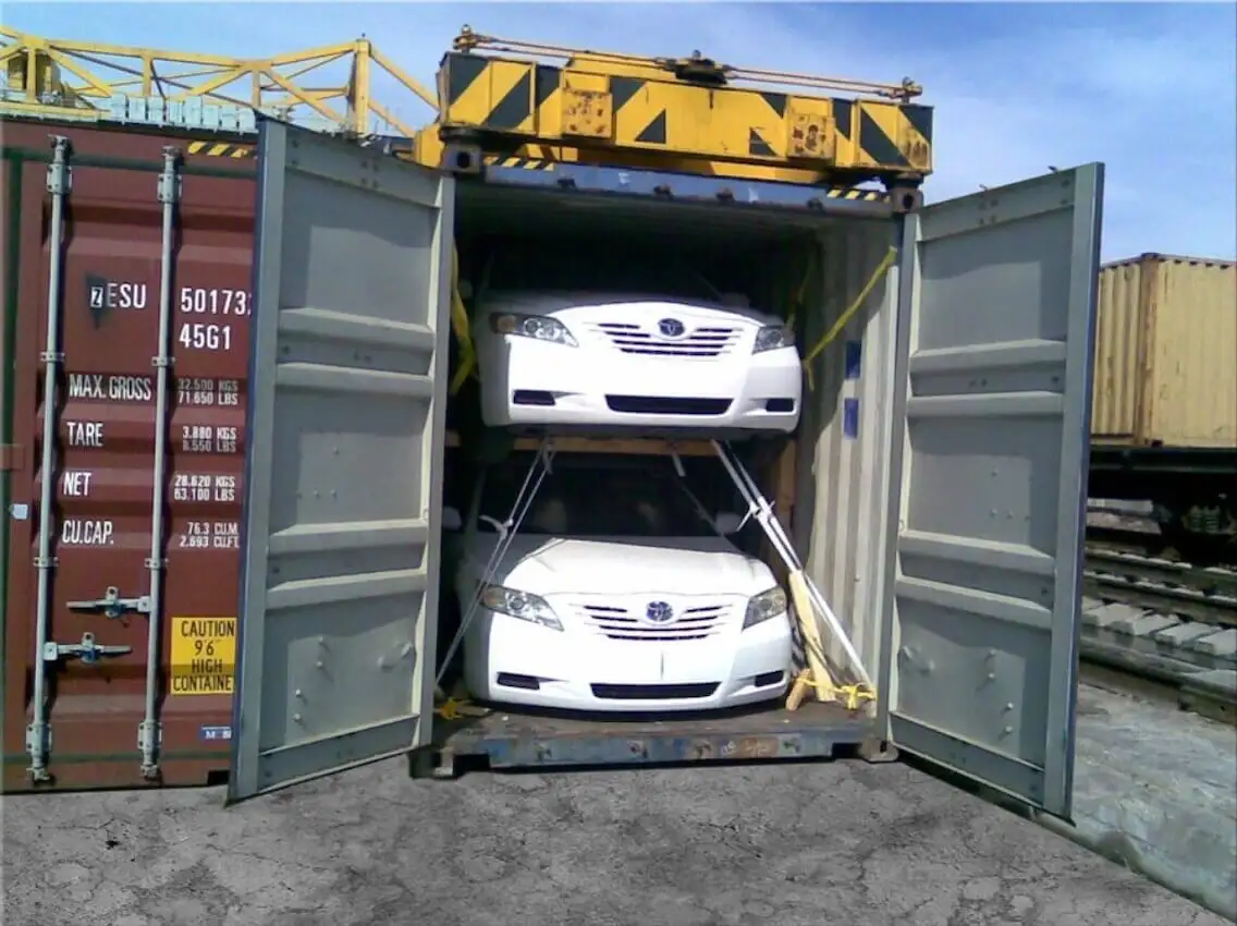 Two white sedans secured by specialized steel racks inside an open shipping container for cost-effective vehicle export.