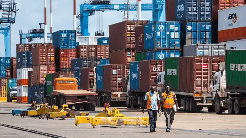 Two port workers walking past a busy cargo terminal full of stacked multi-colored shipping containers and delivery trucks.