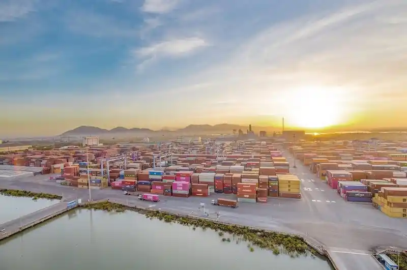Vast aerial view of a huge cargo port at sunrise, showing hundreds of stacked multi-colored shipping containers.