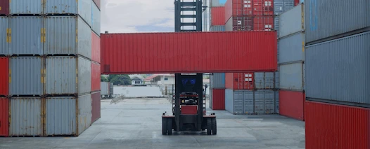 A low-angle view of a reach stacker (heavy forklift) lifting a long red shipping container in a narrow alley between towering stacks of gray and red containers.