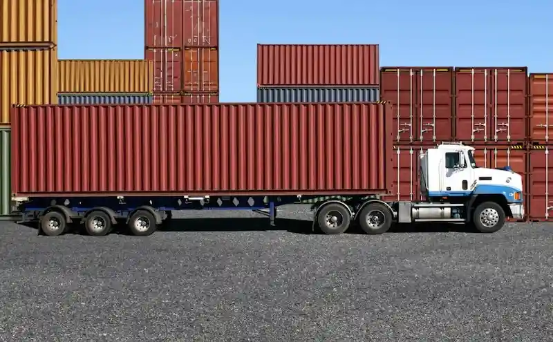 Semi-truck with a flatbed trailer hauling a long red shipping container, with other containers stacked in the background.
