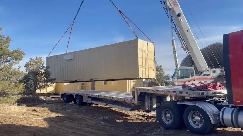 Large crane truck lifting a beige shipping container off its flatbed trailer for precise placement on a rural property.