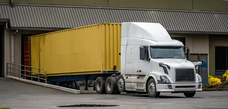 Close-up of a white semi-truck and yellow 40ft container being backed up a ramp for final delivery at a commercial warehouse.