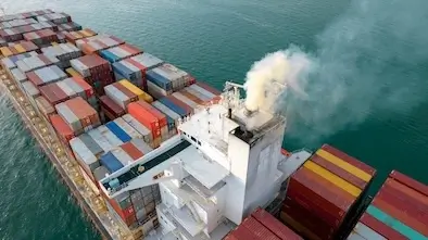 Dramatic aerial view looking down onto the deck of a massive cargo ship loaded with hundreds of multi-colored shipping containers, steaming through blue water.