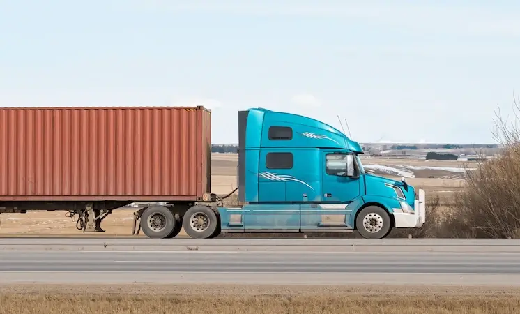 Side view of a bright blue semi-truck hauling a large red shipping container down a highway through open country, illustrating long-haul transport.