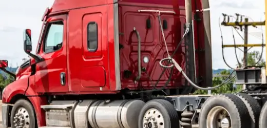 Close-up side view of a powerful red semi-truck cab, highlighting the heavy-duty engine, double axle, and hitch mechanism used for hauling shipping containers.