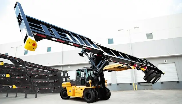 A heavy-duty yellow forklift (reach stacker) lifting a black intermodal shipping container chassis trailer by the corner fittings in an industrial yard