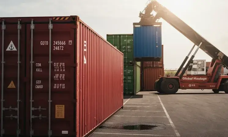 A red shipping container in close-up showing its ID, weight, and specs, with a reach stacker in the background lifting a blue container in a busy terminal.