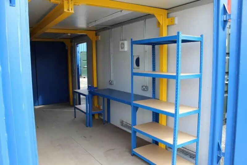 Interior of a modified shipping container workshop showing blue shelving, a workbench, electrical outlets, and yellow structural beams.