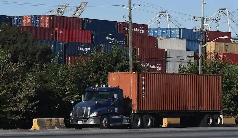 Blue semi-truck hauling a large orange container, passing dense stacks of multi-colored containers and cranes at a port.